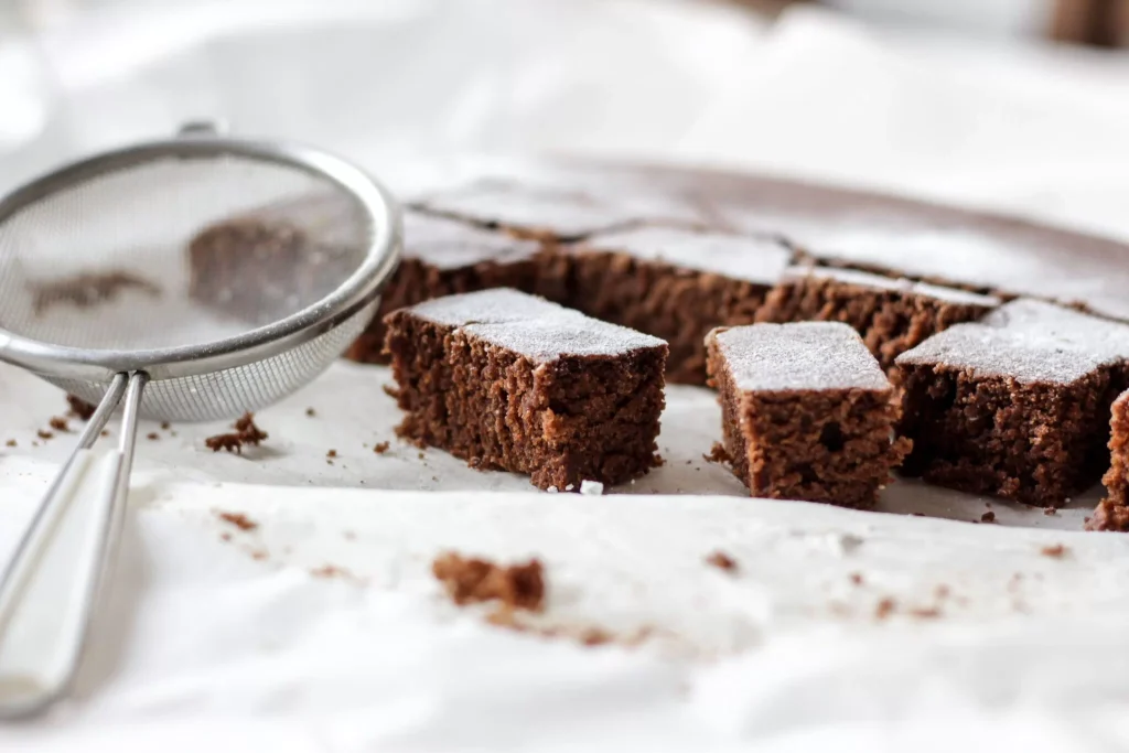 Frisch gebackene Cannabis-Brownies, in Stücke geschnitten und mit Puderzucker bestäubt, neben einem Sieb auf Backpapier – Anleitung für selbstgemachte THC-Edibles